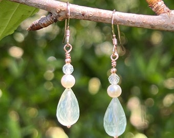 May include: A pair of dangle earrings with a teardrop-shaped green gemstone, a white pearl, and clear crystal beads. The earrings are made with rose gold-toned metal.