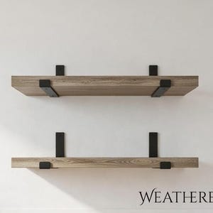 May include: Two horizontal floating shelves made of weathered oak wood with a visible grain pattern. Each shelf is secured to the wall by two black metal brackets that wrap around the front edge. The text WEATHERED OAK is printed below.