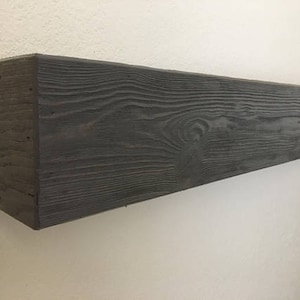 May include: A rectangular floating shelf made of dark stained wood with a visible grain pattern. The shelf features a deep charcoal finish and a textured surface, mounted horizontally against a plain, light-colored wall surface.