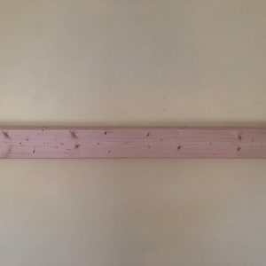 K&ouml;nnte beinhalten: Ein langes, rechteckiges h&ouml;lzernes Wandregal mit einer hellen, rosabeigen Oberfl&auml;che. Die Holzmaserung ist auf der Vorderseite sichtbar und weist mehrere kleine, dunkelbraune Astl&ouml;cher auf, die horizontal &uuml;ber die L&auml;nge des Brettes verteilt sind.