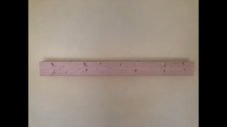 K&ouml;nnte beinhalten: Ein langes, rechteckiges h&ouml;lzernes Wandregal mit einer hellen, rosabeigen Oberfl&auml;che. Die Holzmaserung ist auf der Vorderseite sichtbar und weist mehrere kleine, dunkelbraune Astl&ouml;cher auf, die horizontal &uuml;ber die L&auml;nge des Brettes verteilt sind.