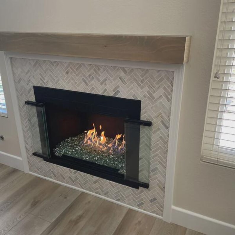 Puede incluir: Una chimenea con marco de metal negro, paneles laterales de vidrio y un lecho de cuentas de fuego de vidrio verde reflectante. El marco est&aacute; revestido con azulejos en patr&oacute;n de espiga claro y rematado con una repisa de madera natural.