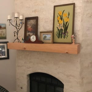May include: A natural light-colored wooden fireplace mantel shelf mounted on a textured stone wall. The mantel holds a black metal candelabra, a dark wood mantel clock, two framed landscape paintings, and a framed floral needlepoint art piece.