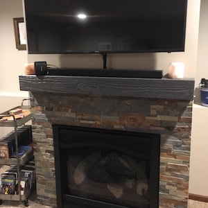May include: A fireplace with a stacked stone facade in shades of gray, tan, and brown, topped with a dark gray wooden mantel. A black television is mounted above the mantel, with a black soundbar and two small light-colored lamps placed below it.