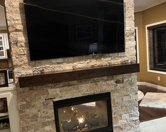 May include: A large rectangular flat screen television mounted above a dark brown wooden mantelpiece. Below the mantel is a black fireplace insert with a glass front, set into a wall constructed of light tan, textured, stacked stone veneer.