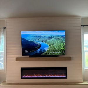 Puede incluir: Un televisor de pantalla plana montado en la pared que muestra una imagen de paisaje de un r&iacute;o serpenteando a trav&eacute;s de colinas verdes, colocado sobre una repisa de madera natural y una chimenea el&eacute;ctrica rectangular con llamas naranjas sobre una pared de listones blancos.