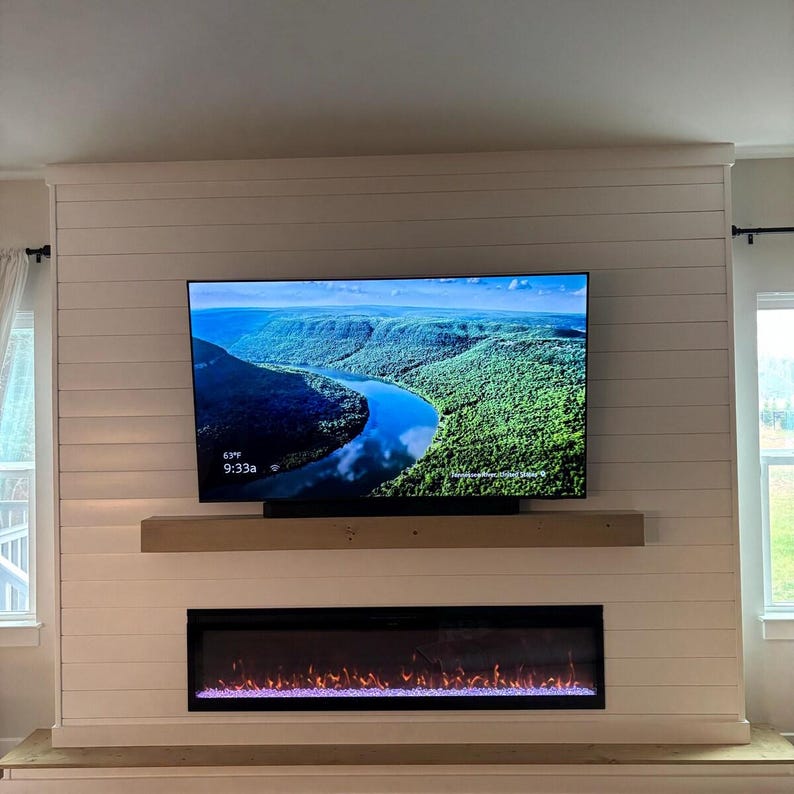Puede incluir: Un televisor de pantalla plana montado en la pared que muestra una imagen de paisaje de un r&iacute;o serpenteando a trav&eacute;s de colinas verdes, colocado sobre una repisa de madera natural y una chimenea el&eacute;ctrica rectangular con llamas naranjas sobre una pared de listones blancos.