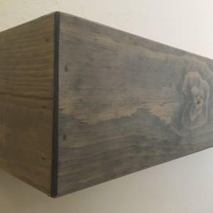 Puede incluir: Un estante de pared flotante rectangular hecho de madera con un acabado te&ntilde;ido en gris oscuro. El estante presenta un patr&oacute;n de veta de madera prominente y un nudo visible en la cara frontal, con bordes oscuros donde se unen los paneles.