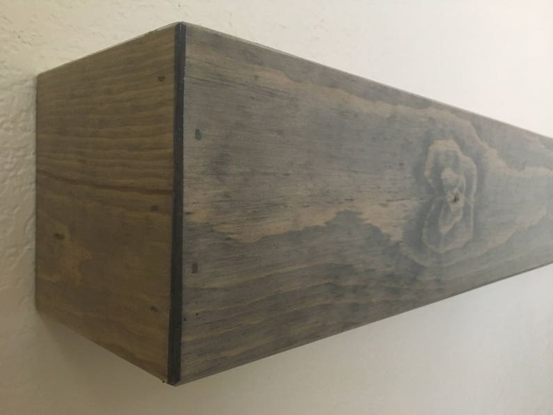 Puede incluir: Un estante de pared flotante rectangular hecho de madera con un acabado te&ntilde;ido en gris oscuro. El estante presenta un patr&oacute;n de veta de madera prominente y un nudo visible en la cara frontal, con bordes oscuros donde se unen los paneles.