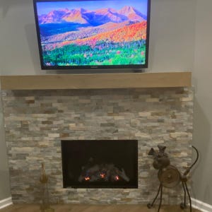 Puede incluir: Una chimenea con un revestimiento de piedra apilada en tonos gris y beige, rematada por una repisa de madera horizontal. Sobre la repisa, un televisor muestra una imagen de paisaje de monta&ntilde;as y &aacute;rboles. Una escultura de metal se sit&uacute;a a la derecha.