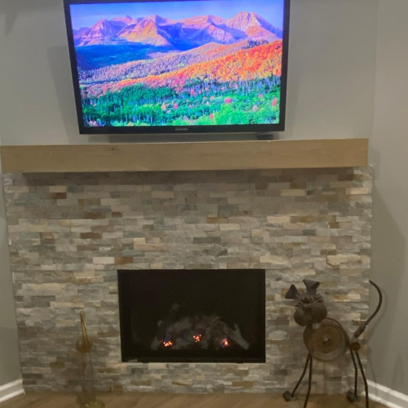 Puede incluir: Una chimenea con un revestimiento de piedra apilada en tonos gris y beige, rematada por una repisa de madera horizontal. Sobre la repisa, un televisor muestra una imagen de paisaje de monta&ntilde;as y &aacute;rboles. Una escultura de metal se sit&uacute;a a la derecha.