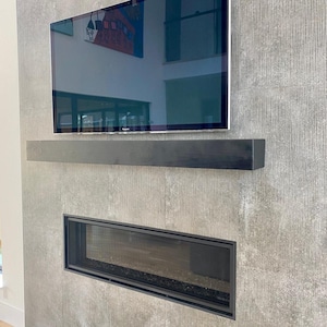 May include: A rectangular black wooden mantel shelf mounted on a textured gray vertical wall surface. The mantel is positioned directly below a flat screen television and above a horizontal rectangular fireplace with a black metal frame.