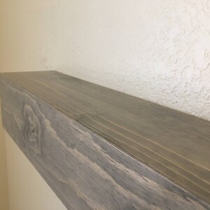 Puede incluir: Un estante flotante rectangular de madera con un acabado liso te&ntilde;ido de gris. La veta de la madera es visible en las superficies superior y frontal, con un nudo natural prominente en el lado izquierdo del panel frontal. El estante est&aacute; montado horizontalmente.