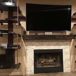 May include: A fireplace with a white stone tile surround and a dark wood mantelpiece. Above the mantel is a large flat-screen television. To the left, a tall shelving unit is constructed from black metal shelves and copper-colored pipes.