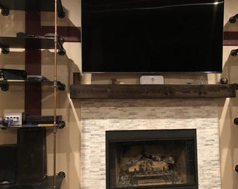 May include: A fireplace with a white stone tile surround and a dark wood mantelpiece. Above the mantel is a large flat-screen television. To the left, a tall shelving unit is constructed from black metal shelves and copper-colored pipes.