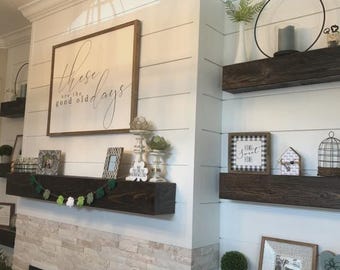 May include: A white shiplap wall featuring a large rectangular sign with the text these are the good old days, dark wood floating shelves, and various decorative items including a green felt shamrock garland, picture frames, and plants.