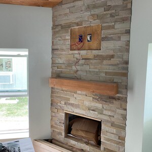 May include: A fireplace surround constructed from stacked, multi-tonal beige and tan stone veneer. A rectangular wooden mantel is mounted horizontally across the center. An open rectangular cavity is at the base, and a recessed panel with exposed electrical wiring is positioned above the mantel.