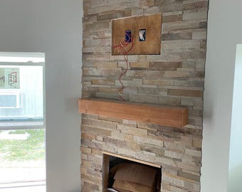 May include: A fireplace surround constructed from stacked, multi-tonal beige and tan stone veneer. A rectangular wooden mantel is mounted horizontally across the center. An open rectangular cavity is at the base, and a recessed panel with exposed electrical wiring is positioned above the mantel.