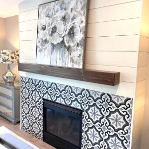 May include: A fireplace surround featuring a dark brown wooden mantel, a black firebox, and decorative square tiles with a black, white, and gray geometric pattern. Above the mantel hangs a square canvas painting of white flowers.