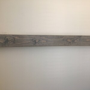 Puede incluir: Un estante flotante rectangular y alargado hecho de madera con un acabado en tono gris. La superficie muestra un patr&oacute;n de veta de madera natural visible con nudos y vetas m&aacute;s oscuras que recorren horizontalmente toda la pieza.