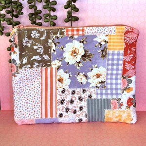 Può includere: Una pochette rettangolare con cerniera caratterizzata da un design patchwork con vari motivi in tessuto, tra cui stampe floreali in viola, bianco e marrone, strisce arancioni e bianche, quadretti vichy arancioni e gessato grigio, rifinita con una cerniera arancione chiaro.