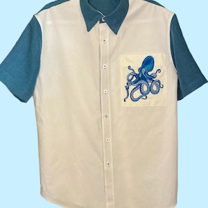Puede incluir: Una camisa de manga corta con botones, panel frontal blanco y mangas y cuello azules. Un bolsillo en el pecho izquierdo muestra una ilustración detallada de un pulpo azul. La camisa cuenta con un cuello estándar y una hilera vertical de botones blancos.