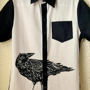 May include: A short-sleeved button-down shirt with a black and white design. The shirt features a large black raven graphic on the front, a black pocket, and black collar and sleeves. The shirt is made of a light-colored fabric.