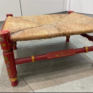 Puede incluir: Un reposapiés rectangular bajo con asiento de fibra natural tejida y estructura de madera pintada de rojo. Las cuatro patas cilíndricas y los travesaños laterales están decorados con motivos florales pintados a mano en amarillo y verde.