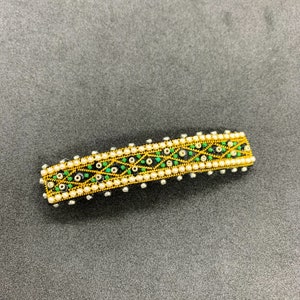 May include: A rectangular hair barrette with a gold-coloured frame. The barrette features a pattern of green and black accents, small white beads, and a gold-coloured border. The barrette is approximately 7.6 cm long.
