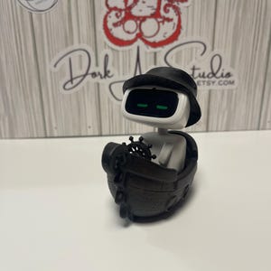 May include: A white and black robot figurine wearing a black hat and sitting in a small wooden boat. The robot has green eyes and a steering wheel. The boat is dark brown. The background has a wooden panel design with stickers and text.