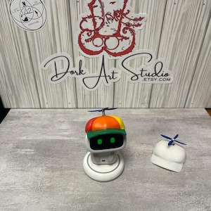 May include: A white robot toy with a colorful hat and propeller, sitting on a white charging base. A second white hat with a propeller is next to it. The text "Dork Art Studio etsy.com" is visible.
