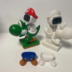 May include: Two white plastic figurines of Mario and Luigi characters riding on green and white Yoshi dinosaurs. The figurines are part of a set of four, with two additional pieces shown in the image: a blue and orange Mario hat and a white Luigi hat.