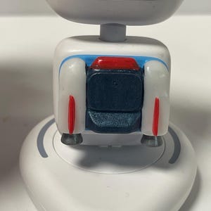 May include: A white robot-like device with a rounded top and a square midsection. The midsection features blue, red, and dark blue accents. The base is white and oval-shaped, with a gray curved line.