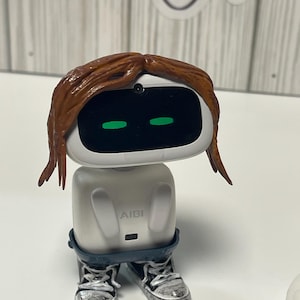May include: A white AI robot figurine with brown hair and green eyes. The robot is wearing blue jeans and silver sneakers. The word "AIBI" is printed on the front of the robot.