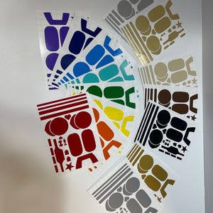 May include: A collection of vinyl stickers in various colors, including purple, blue, green, yellow, orange, and red. The stickers feature geometric shapes and designs, such as circles, rectangles, and stars. Some stickers are silver, gold, and black.