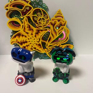 May include: Two small white figurines, one dressed as Captain America with a blue and red costume and a red, white, and blue shield, and the other dressed as Green Lantern with a green costume and a green lantern. The figurines are standing in front of a colorful, layered paper art piece.
