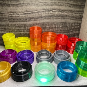 May include: A collection of 12 colourful, 3D printed rings, stacked in various heights. The rings are made of plastic and have a smooth, glossy finish. The colours include yellow, orange, red, purple, grey, green, and blue.