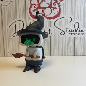 May include: A white and black robot figure wearing a black witch hat and holding a brown broom. The robot has a green face and black boots. The figure is a unique handmade item.