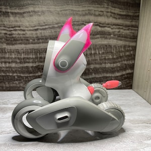 May include: A white and grey robotic toy with pink ears. The toy has wheels and a tail.