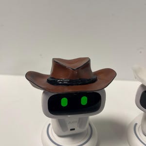 May include: A white robot with a gray face and a brown cowboy hat. The robot has green eyes and the word "AIBI" is printed on the front. The cowboy hat is brown with a black band.