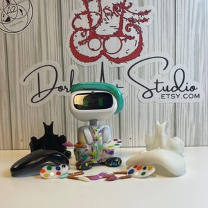 May include: A white and green robot figurine with colorful paint splatters, paint palettes, and paintbrushes. The robot has a green headband and a screen for a face. The image includes stickers with text, including "Dork Studio .Etsy.com".