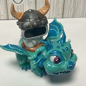 May include: A whimsical, hand-painted ceramic dragon figurine in shades of teal and blue, adorned with a Viking helmet. The dragon has large, expressive eyes with pink accents and small horns. The figurine is a unique decorative item.
