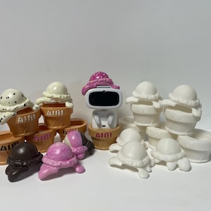 May include: A collection of miniature ice cream cones in various colors and designs, including white, pink, and brown. Some cones have a white base with a pink or brown ice cream scoop on top, while others have a brown base with a white ice cream scoop on top. There is also a small white robot figure with a pink hat.