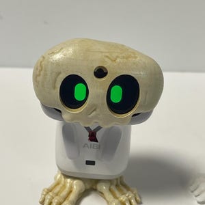 May include: A novelty figurine of a skeleton-like character. It has a beige head, large black eyes with green irises, and a white body. The figurine has skeletal feet and the text "AIBI" on the chest.