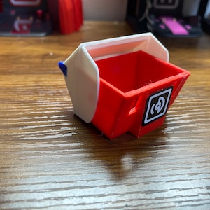 Könnte beinhalten: Eine kleine, rote und weiße Plastik-Aufbewahrungsbox mit einem Scharnierdeckel. Die Box hat einen schwarz-weißen Aufkleber auf der Vorderseite mit dem Buchstaben "D" in einem Kreis.