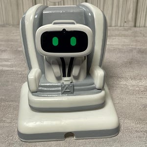 May include: A white and gray robot-themed figurine with a chair-like design. The robot has a rectangular face with two green eye-like lights. The figurine is on a rectangular base and has a modern, minimalist aesthetic.
