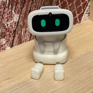 May include: A white robot toy with a rectangular head and two green eye-shaped lights. The robot has small arms and legs, and the word "AIBI" is printed on its body. The robot is sitting on a wooden surface.