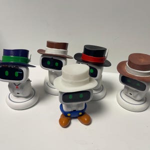 May include: A group of robot figures, each wearing a different hat. The hats are in various colours such as brown, black, white, and blue, some with contrasting bands. The robots have green eyes and are on white bases.