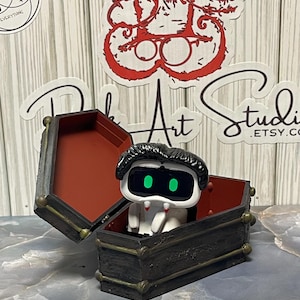 May include: A small, white figurine of a vampire with green eyes and black hair sits inside a miniature coffin. The coffin is black with gold accents and a red interior. The coffin lid is open.