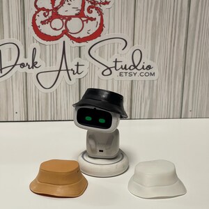 May include: A white robot with green eyes wearing a black bucket hat, with two additional bucket hats in tan and white. The image also includes a sticker that says "Dork Art Studio .etsy.com".